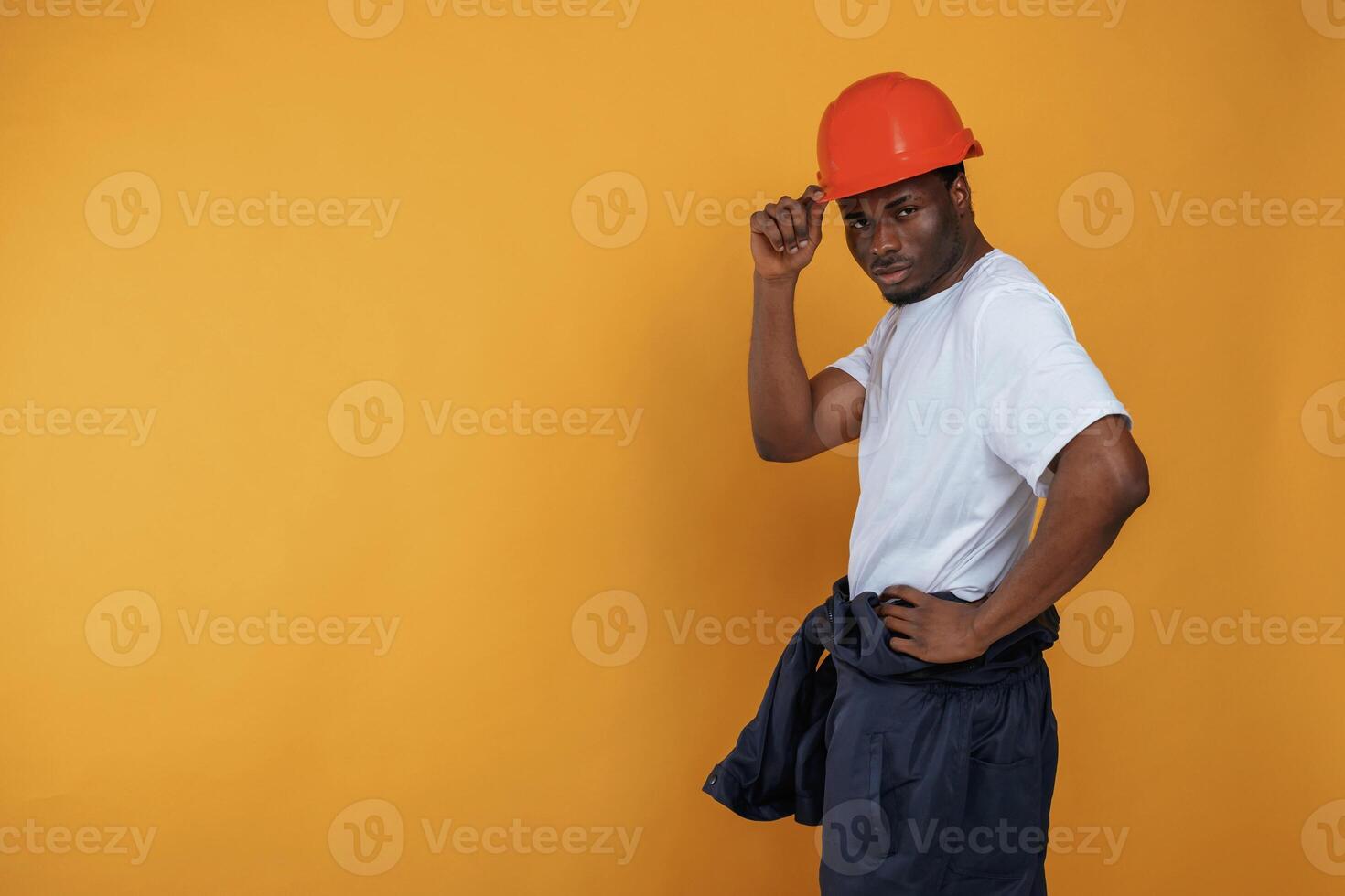 Standing and posing,, construction worker in hard hat. Handsome black man is in the studio against yellow background photo