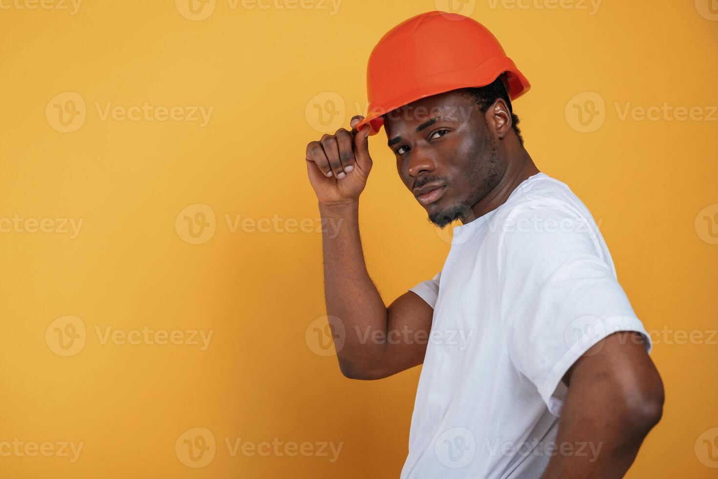 In ,orange ,hard hat. Construction worker. Handsome black man is in the studio against yellow background photo