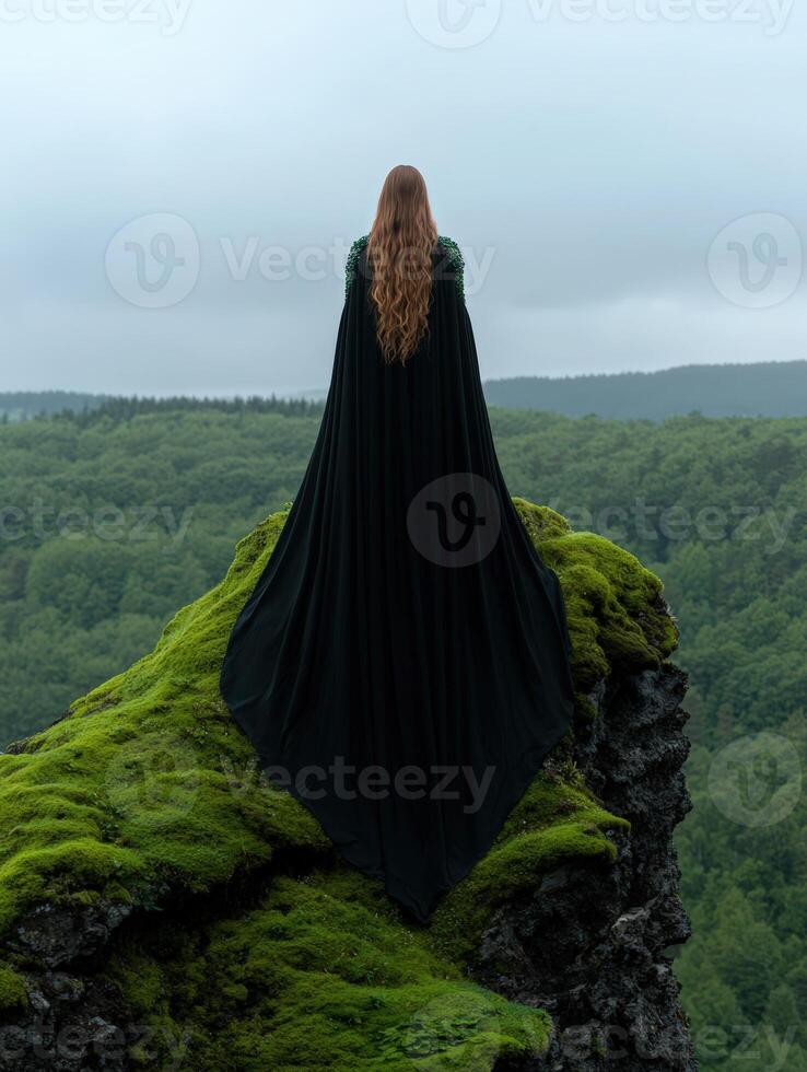 Woman in a long cloak standing on a mossy cliff overlooking a forest landscape photo