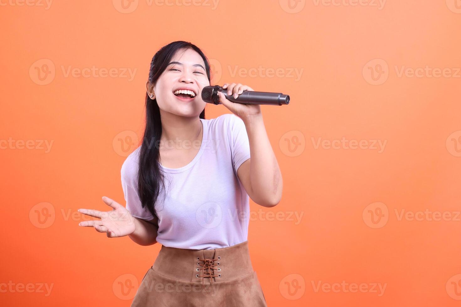 A young Asian woman happily sings into a microphone, her eyes closed, exuding joy and passion for music, isolated on orange background. photo