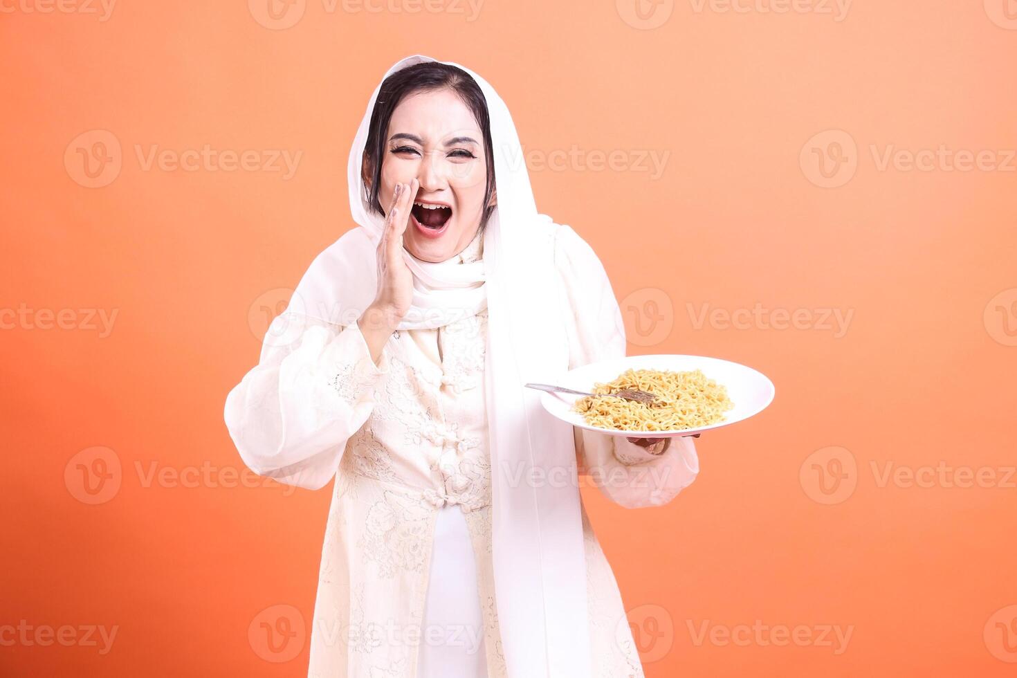 A cheerful Muslim woman wearing a hijab holding a plate of noodles and announcing with joy. isolated on a orange background. photo