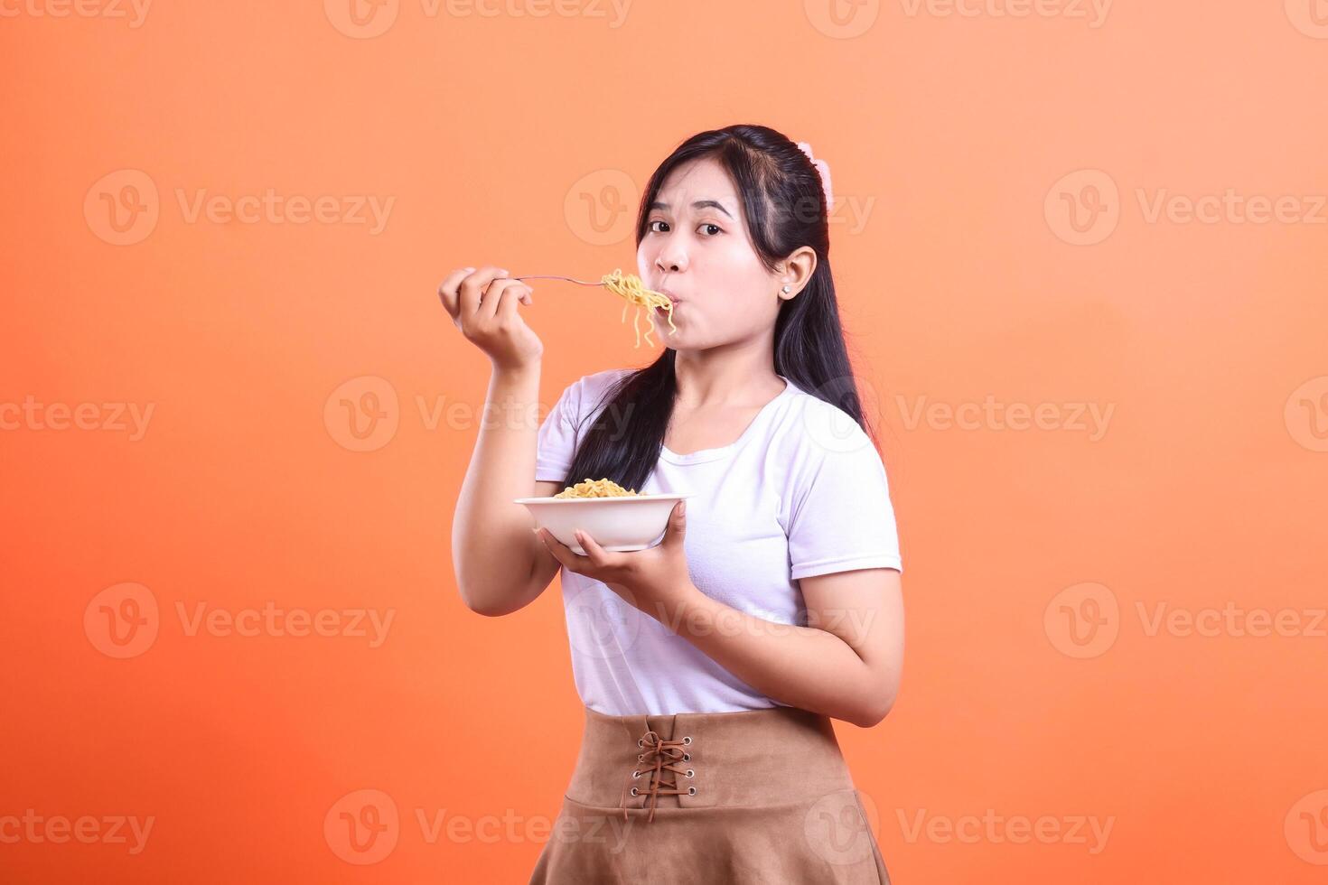 A young woman is enjoying her meal by eating noodles with a fork, she looks happy and pleased with the food, isolated on orange background photo