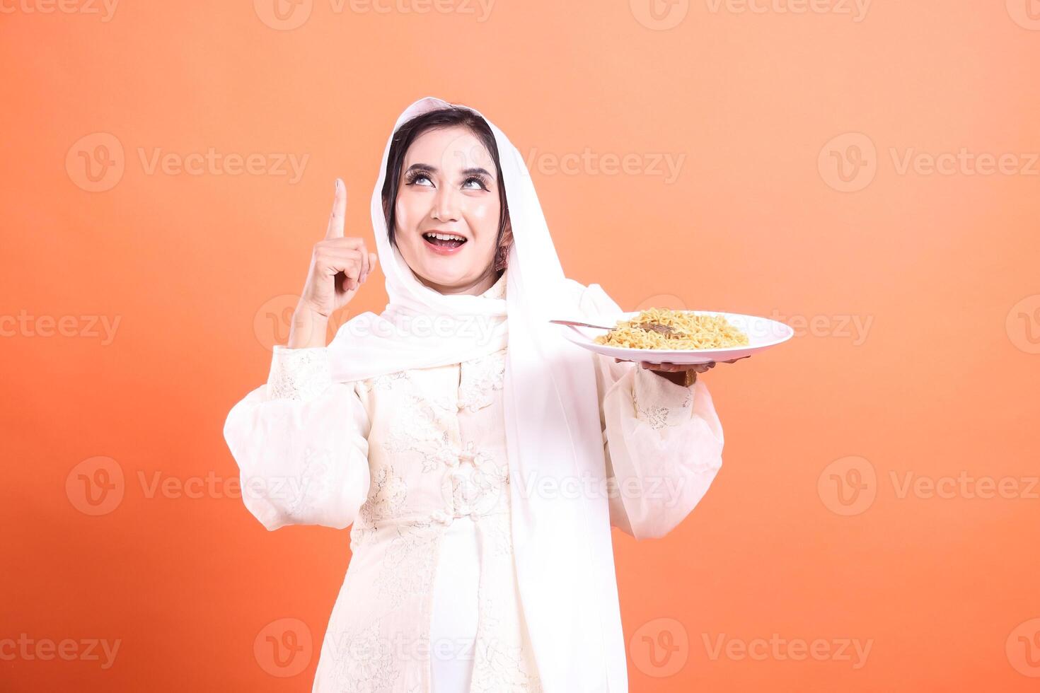 A happy Muslim woman in traditional white clothing holding a plate of noodles and pointing upward. isolated on a orange background photo