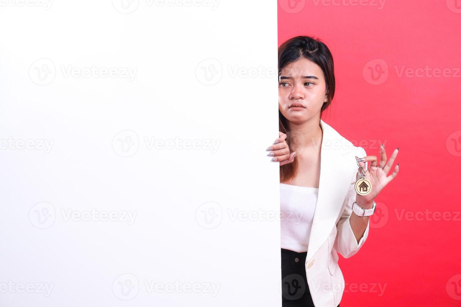A woman with dark hair holds keys with a house charm while partially concealed behind a large white board, isolated on a red background photo
