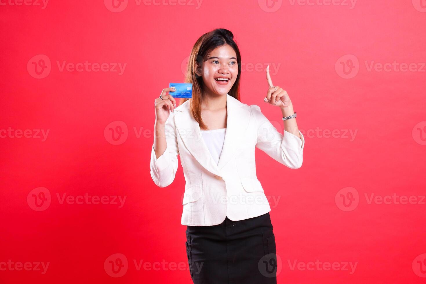 A woman in a white suit holds a credit card and points up with her index finger while smiling, isolated on a red background photo