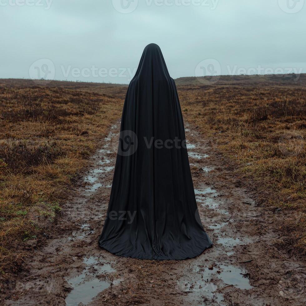 Mysterious figure in black cloak stands on muddy path photo