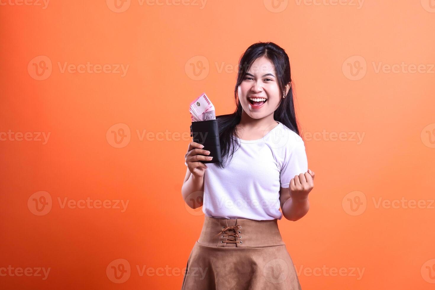 A young woman smiling broadly while holding a black wallet containing rupiah money in her hand isolated on an orange background wearing a white shirt and brown skirt and very happy. photo
