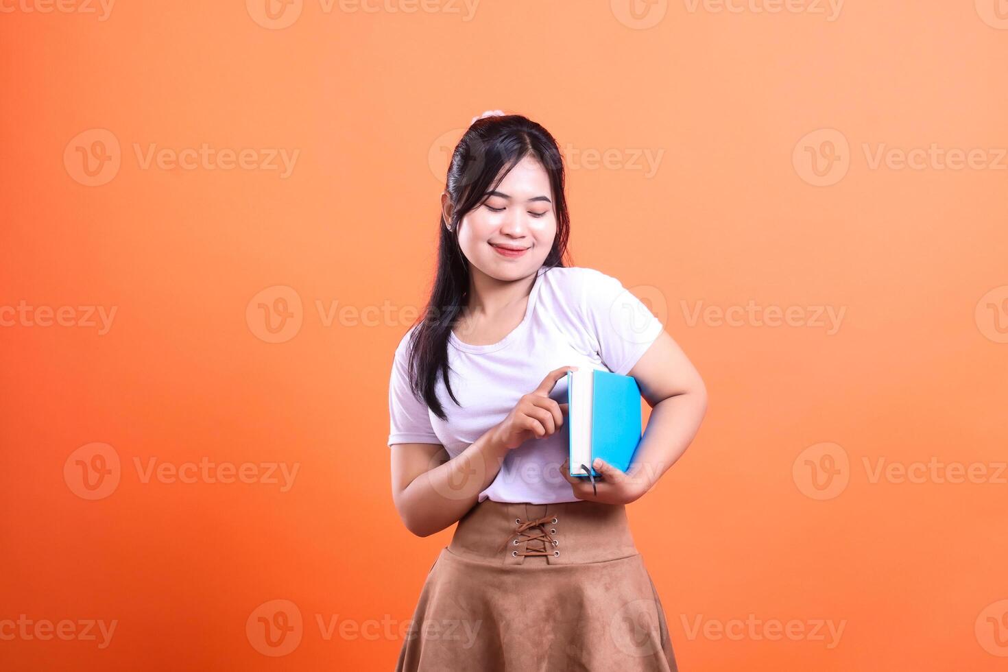 A person in a white shirt and brown skirt holding a book with a solid orange background in studio photo