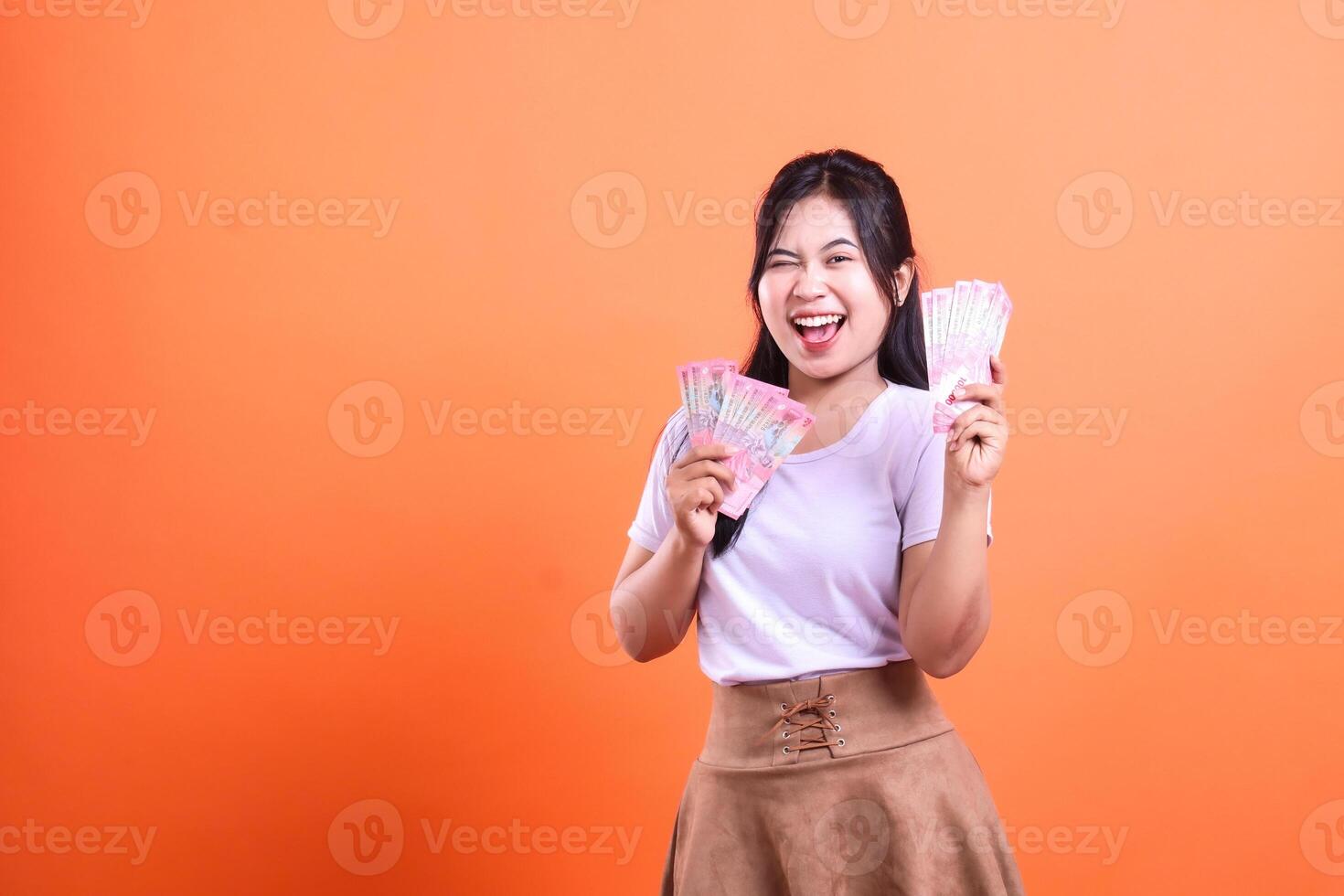 A woman smiles while holding two stacks of Indonesian Rupiah banknotes with a happy expression. The woman is wearing a white top and a brown skirt. isolated on an orange background photo
