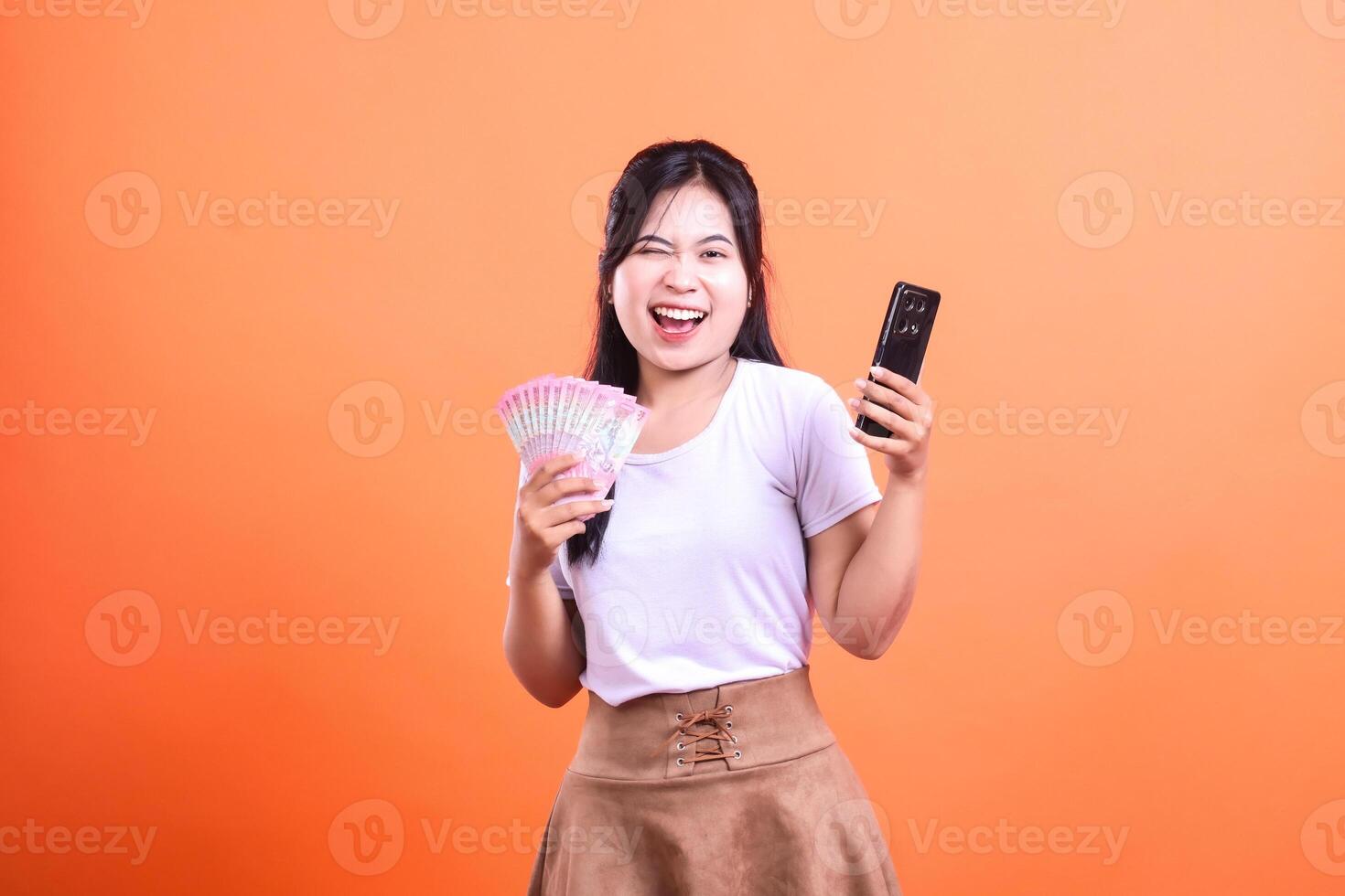 Woman holding money and smart phone device smiles on an orange background for online payment or income photo