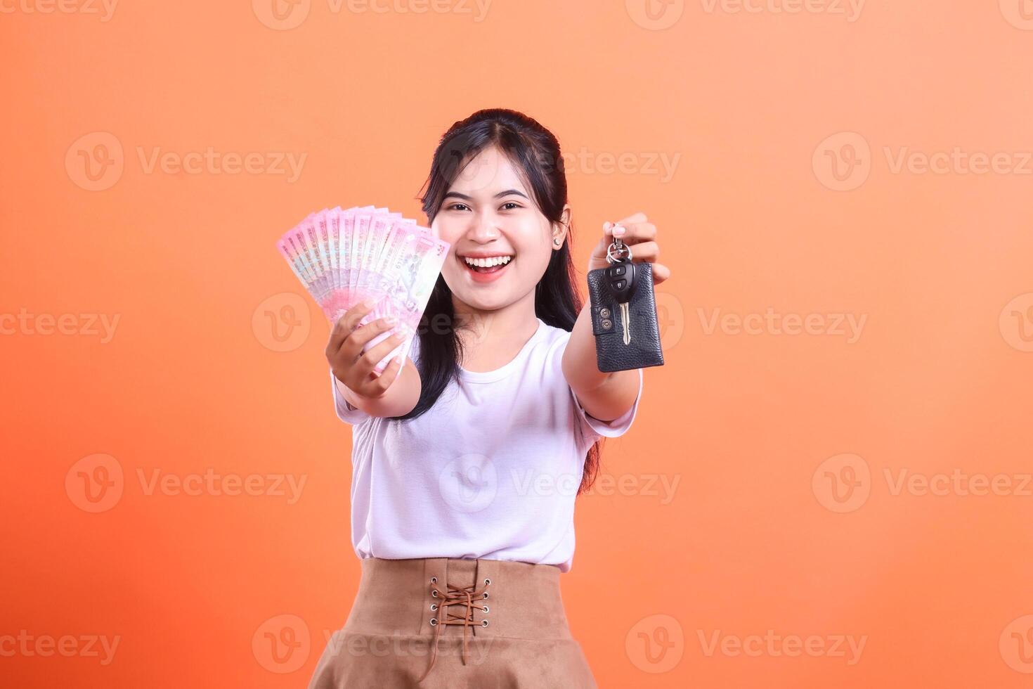 Delighted young Asian woman showcasing a fan of money and car keys with a cheerful smile. isolated on orange background. photo