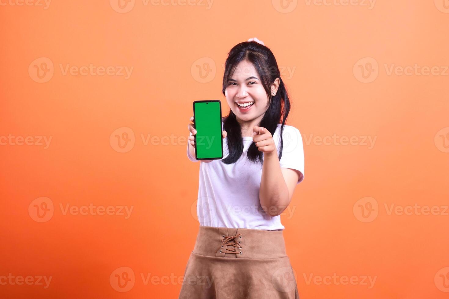 Smiling woman holding a smartphone with green screen, on orange background, and pointing at camera photo