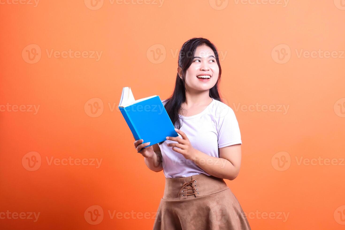 A woman in a white shirt and brown skirt holds a blue book and looks to the side isolated on orange background photo