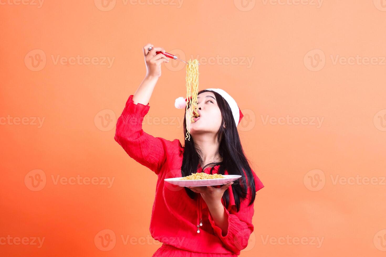 gesture of female wearing red christmas blouse, Santa hat, slurping noodles upwards look left, hand holding fork filled with noodles above head and white plate filled with noodles, isolated photo