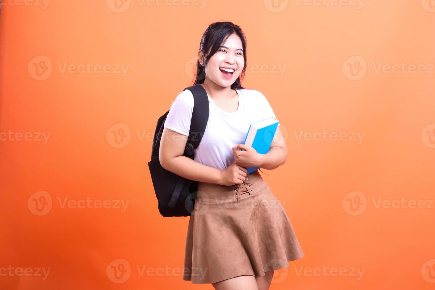 Smiling female student in white shirt, brown skirt, and black backpack holding a blue book on orange background photo
