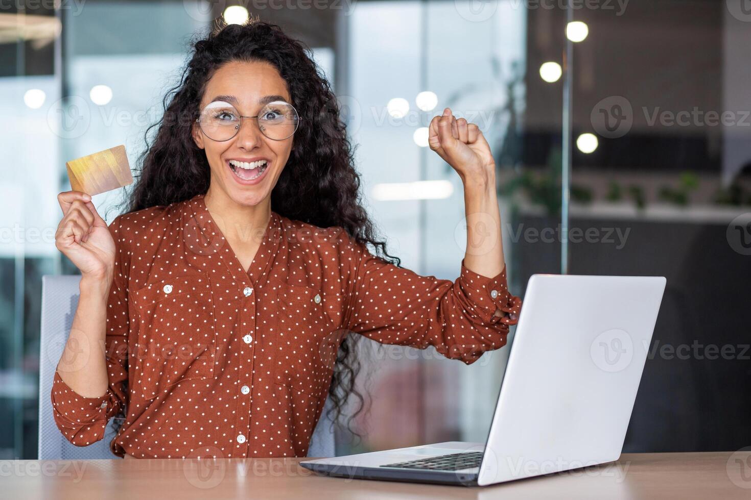 Hispanic attractive woman is delighted with the purchase she made online shopping. Curly brunette in glasses sits at table in front of laptop, holds card in her hands and happily makes online payment. photo