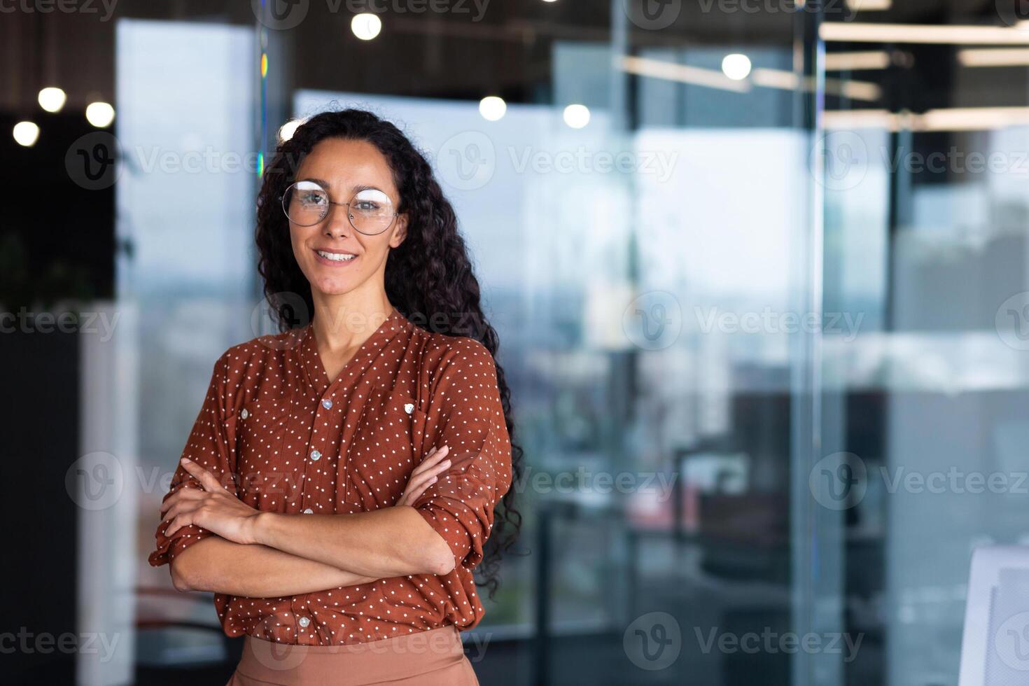 Portrait of happy and successful business woman, boss in shirt smiling and looking at camera inside office with crossed arms, Hispanic woman with curly hair in corridor. photo