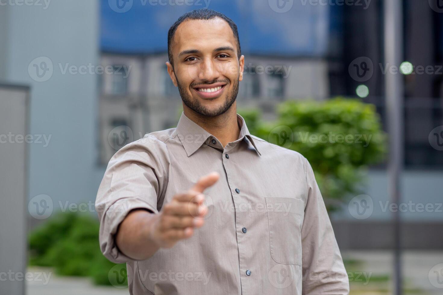 Portrait of a successful and smiling young businessman, hispanic man standing outside an office building and extending his hand to the camera in greeting. photo