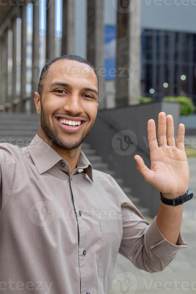 Hispanic smiling young man standing near office building and talking on camera phone, greeting and waving. Vertical photo. photo