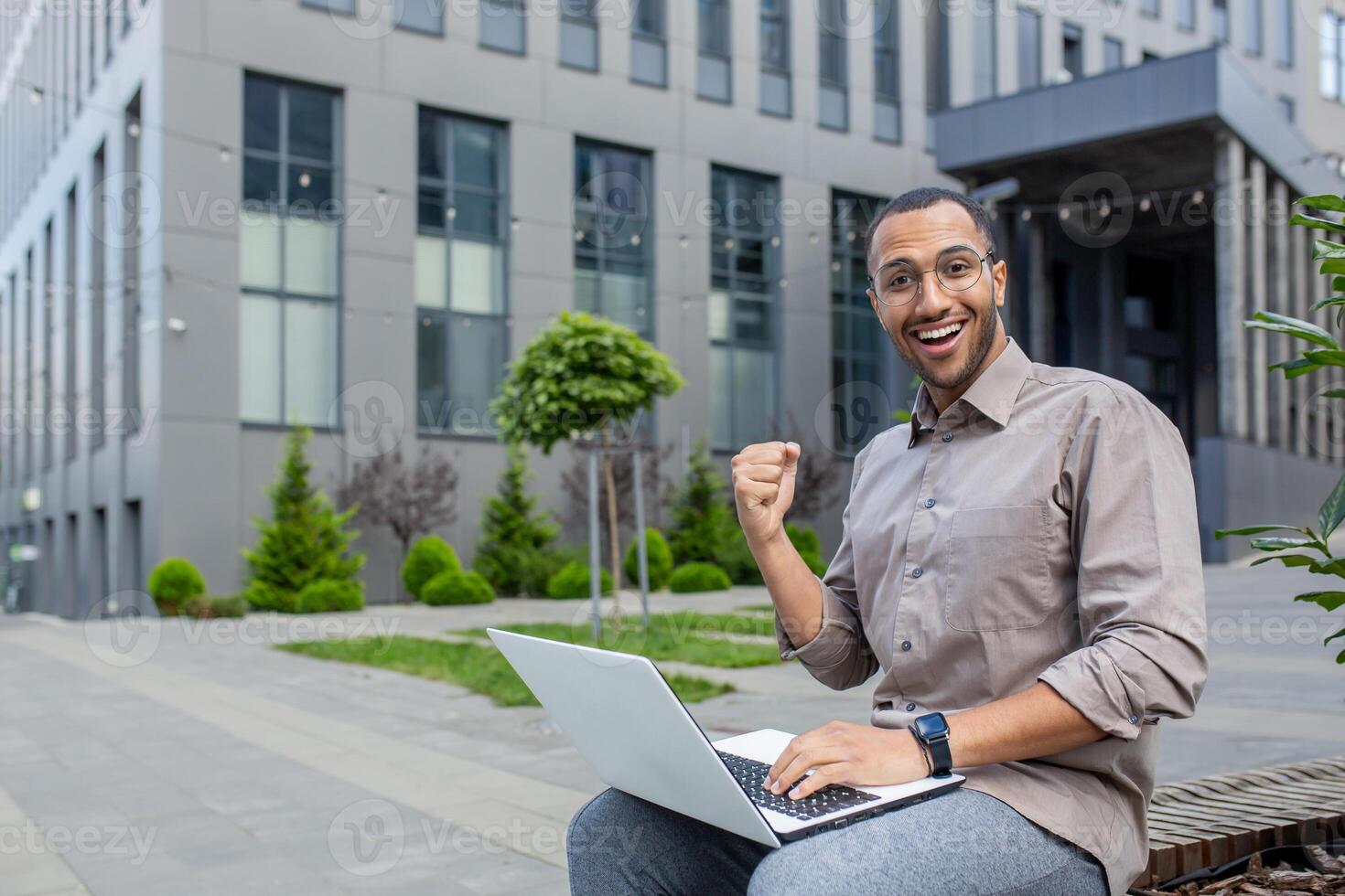 Portrait of a young hispanic male student sitting with a laptop on a bench outside the campus, looking forward to the camera with an achievement and result showing a success and victory gesture. photo