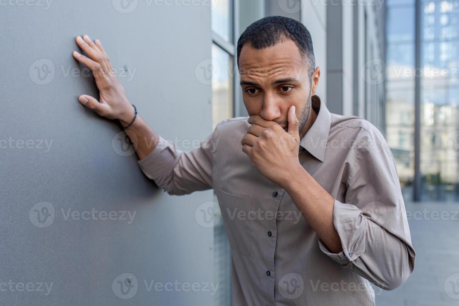 Sick arab male covering mouth with hand while heaving nausea and leaning on concrete wall outdoors. Stressed man trying preventing vomiting after getting gastrointestinal virus from spoiled lunch. photo