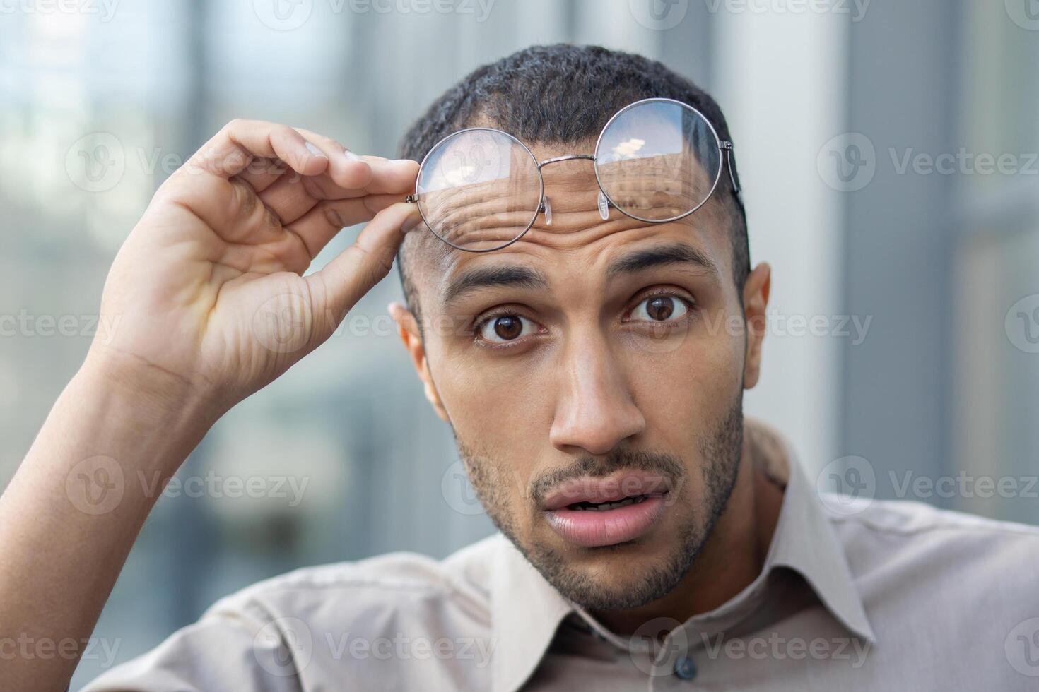 Portrait of surprised arab man looking amazed at camera and taking off round eyeglasses on blurred background. Shocked unshaved guy hearing breaking news or getting unexpected offer outdoors. photo