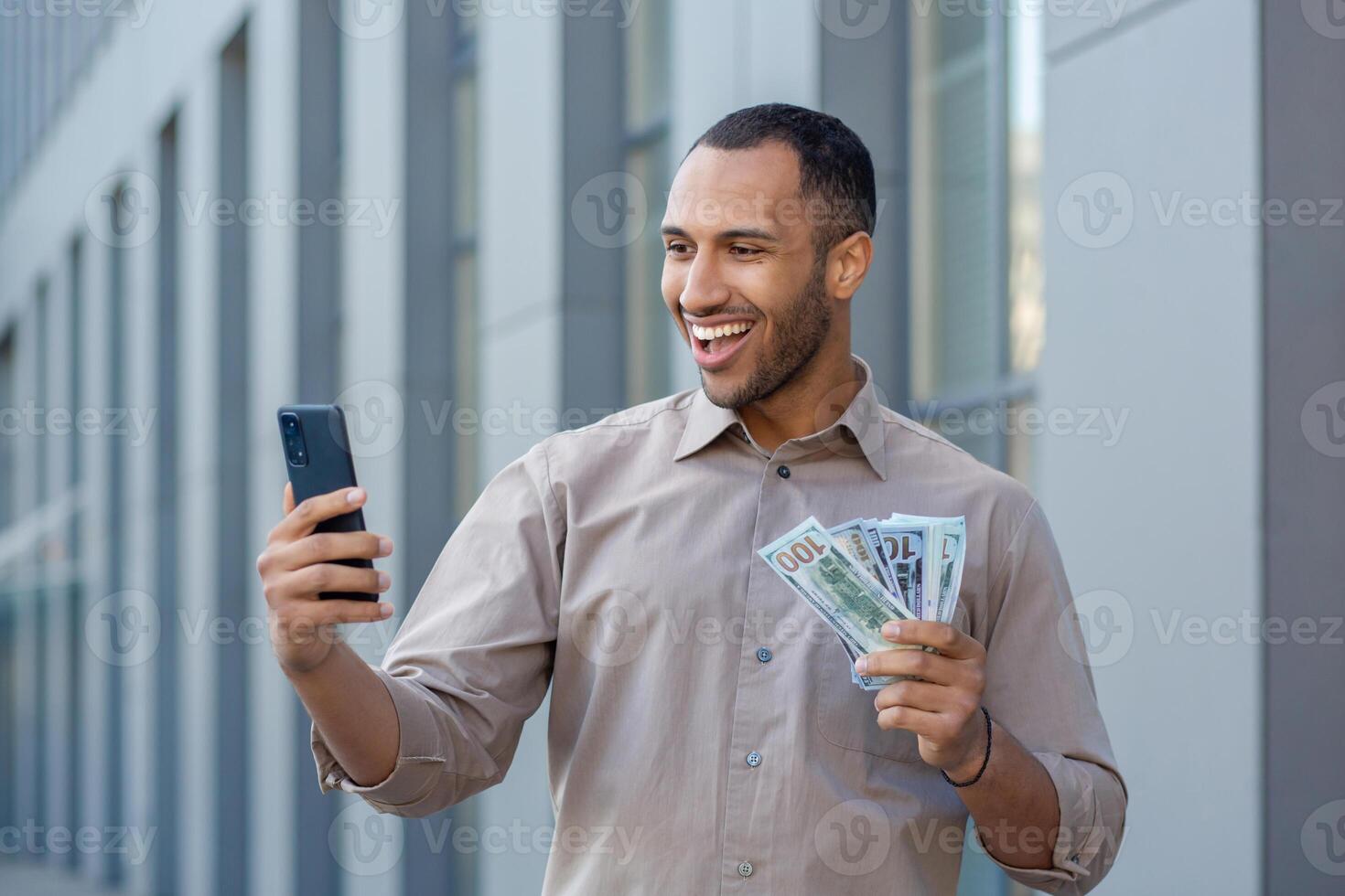 A happy hispanic man is standing on the street near a business center, holding cash money in his hands and talking on a call on a mobile phone camera. photo