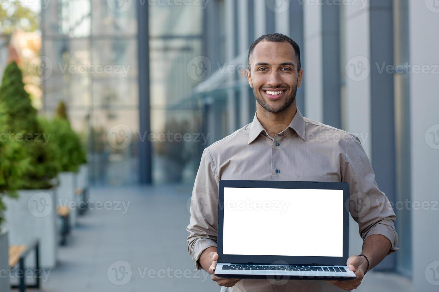 Portrait of a smiling hispanic man holding a mockup of a laptop with a white screen, standing on the street near an office building and showing the device to the camera. photo