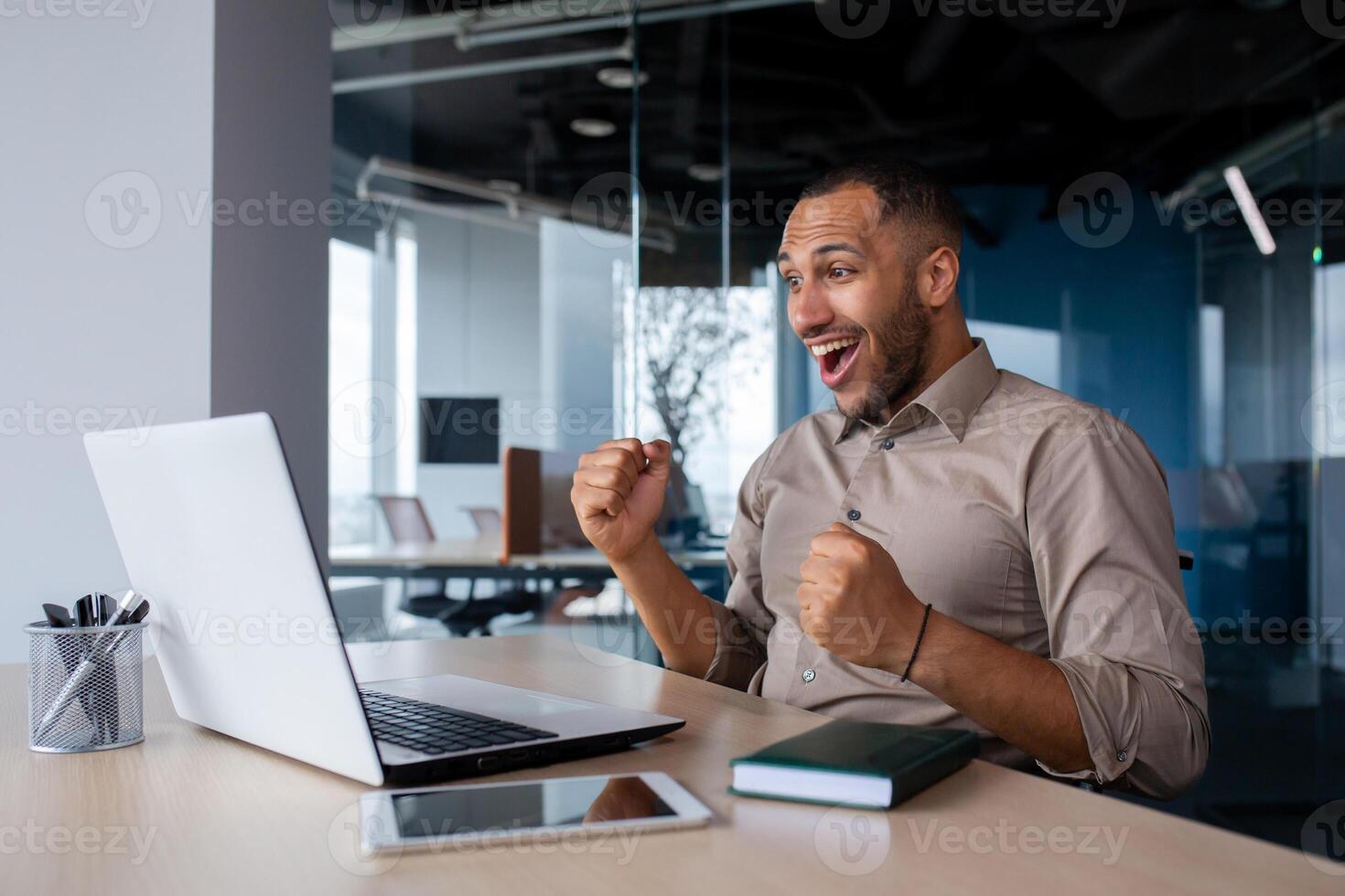 Successful businessman celebrating successful completion of project, man holding hands raised received online notification of triumph and good achievement results on laptop, working inside office. photo