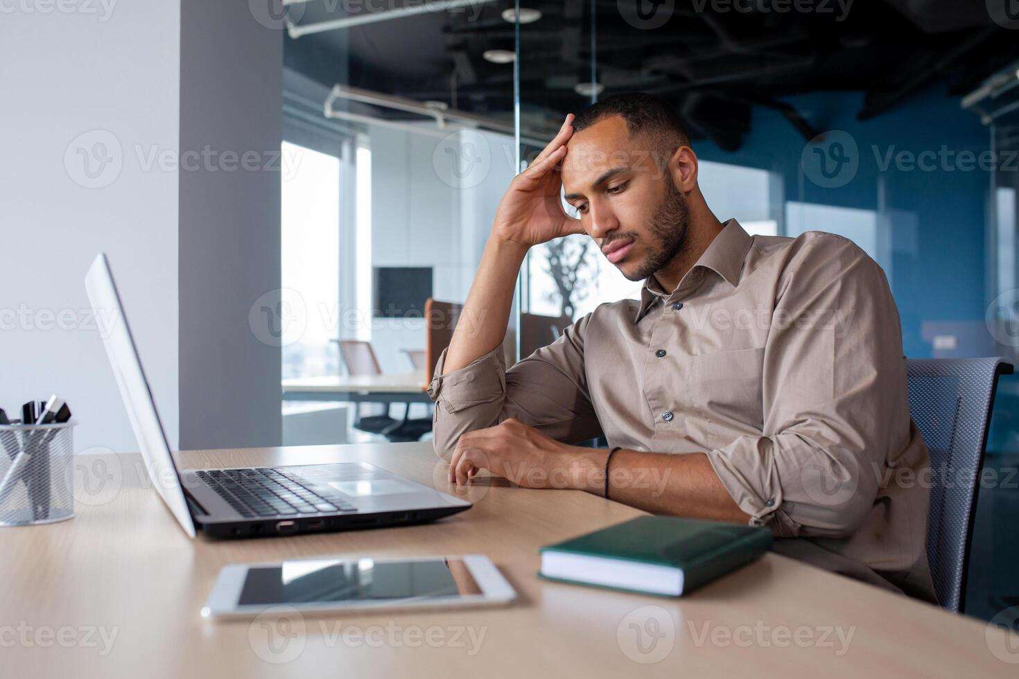 Upset tired young african american man sitting at desk with laptop in office, tired of work holding hand on head, suffering from headache, bored in office. photo