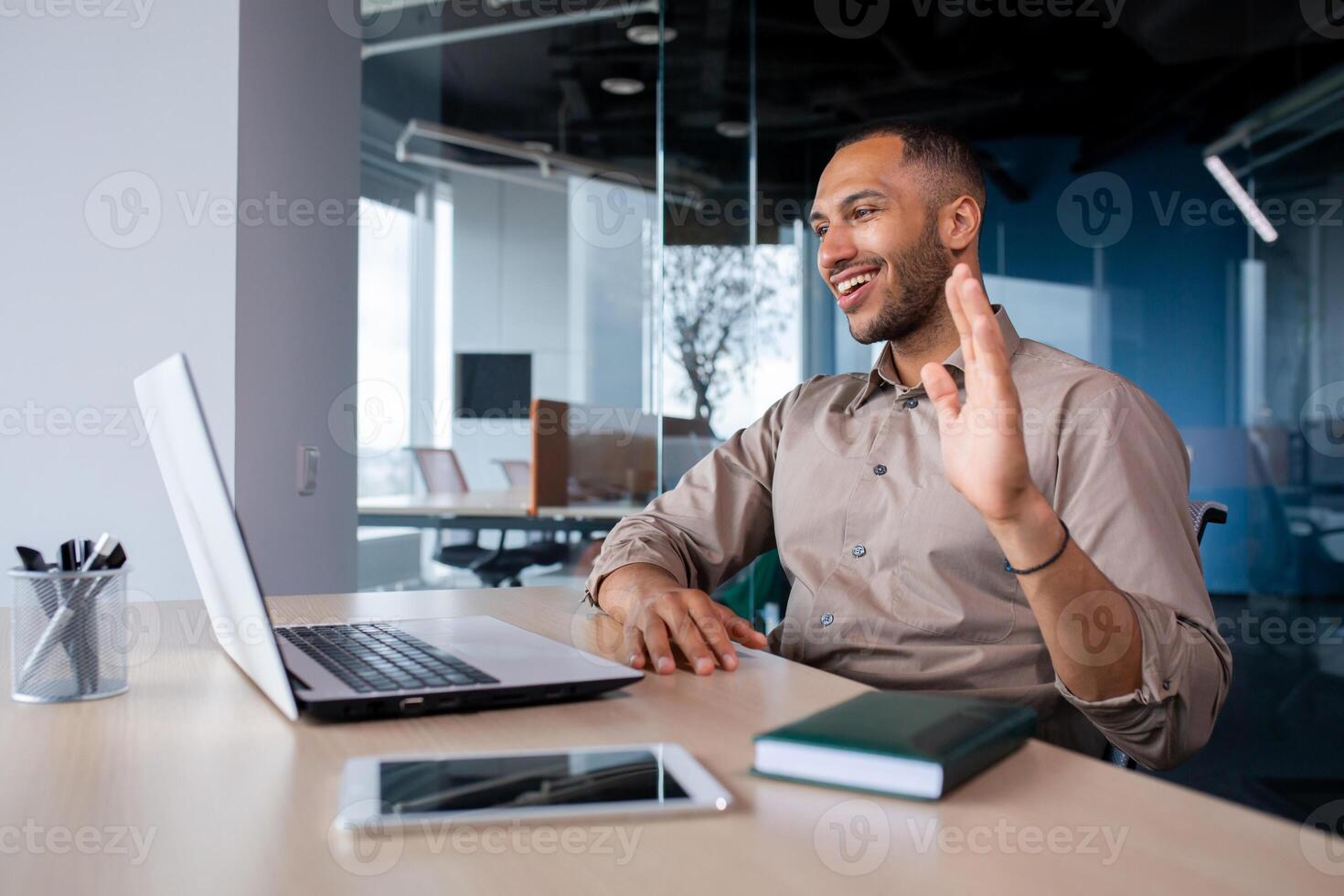 Happy successful and satisfied man talking on call using laptop inside office, businessman waving hand smiling and greeting colleagues and partners, remote online meeting presentation. photo