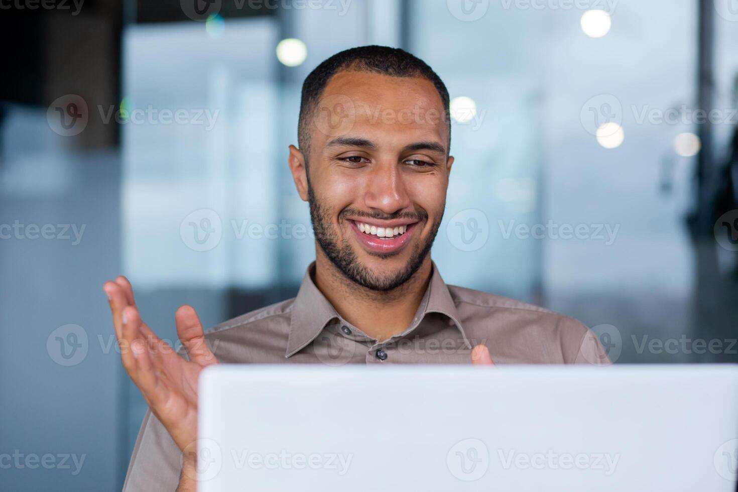 Close-up of a happy successful hispanic businessman making a call from a laptop in a modern office, a young programmer is happy about a successful job, smiling at the computer screen. photo