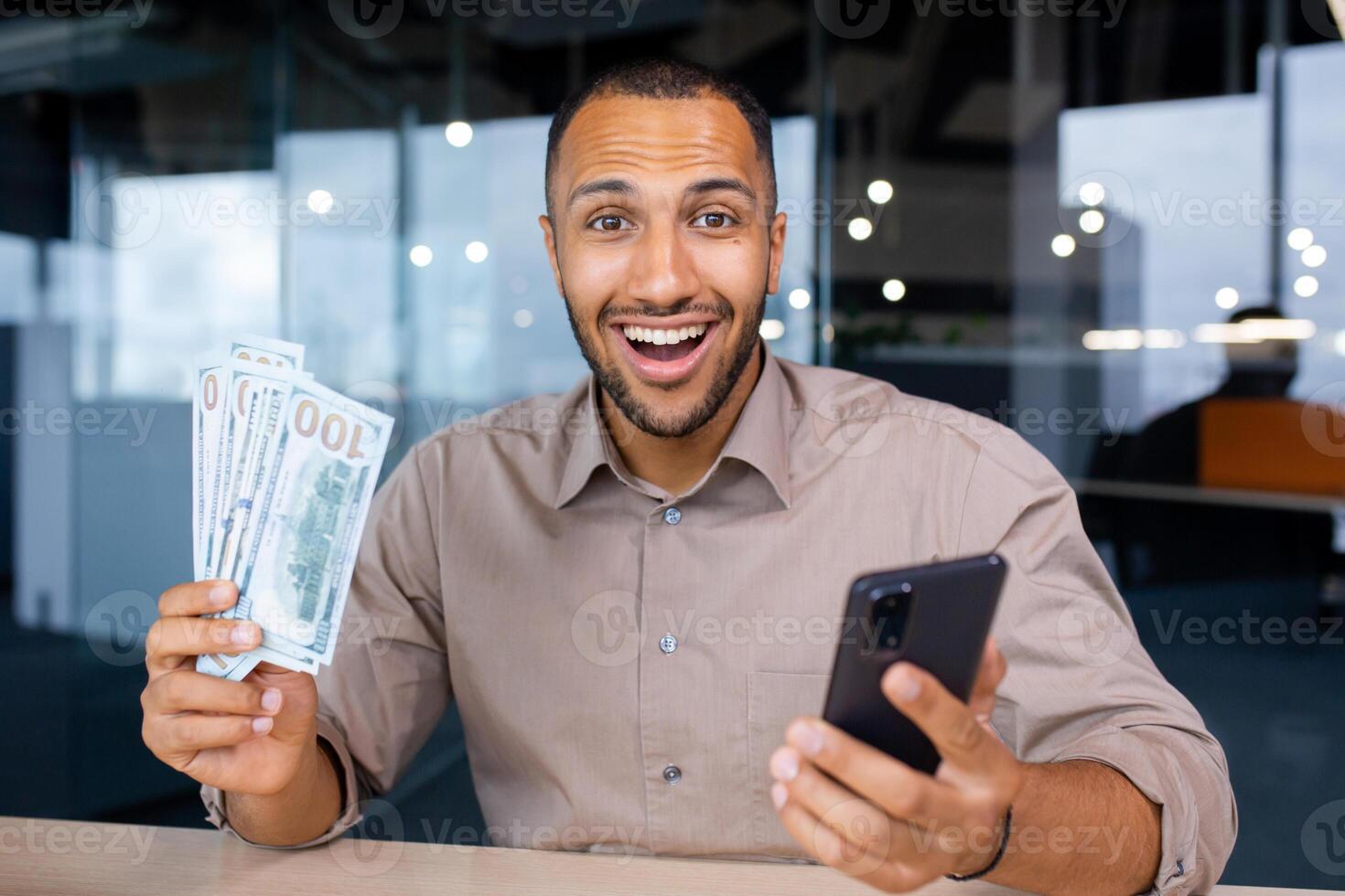 Excited man sitting in modern office holding smartphone and wad of dollars in hands, happy with winning, smiling and looking at camera. photo