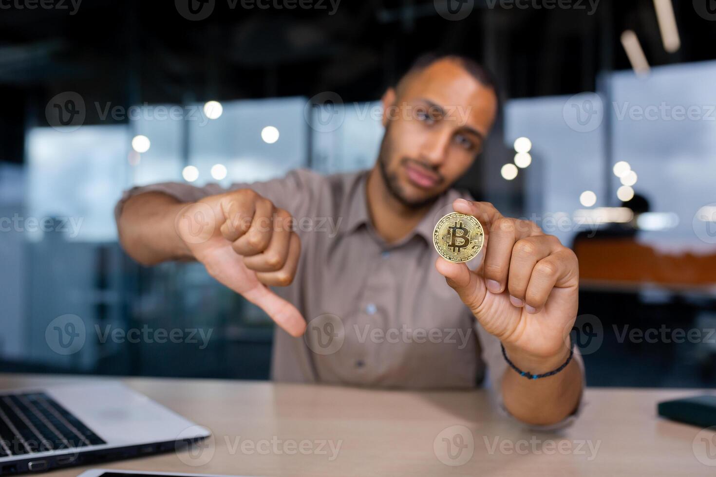 Young man sitting in office with sad face holding bitcoin virtual currency in hands, showing dislike thumb down, rejection concept. photo