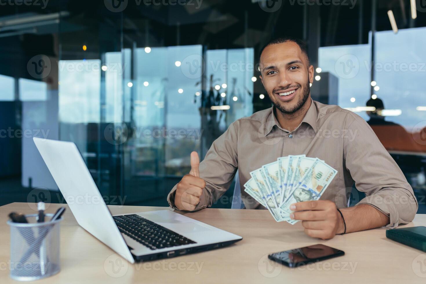 Portrait of successful young financier investor, man smiling and looking at camera, working inside modern office building, holding dollar money cash in hands and showing positive thumbs up photo
