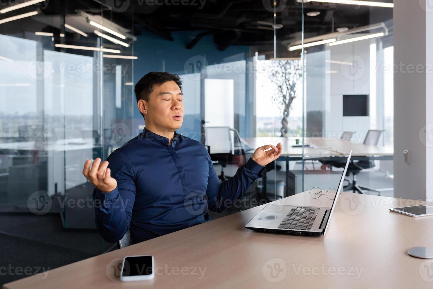 Asian businessman meditating in office, man working at desk using laptop, worker with closed eyes in lotus pose inside building. photo