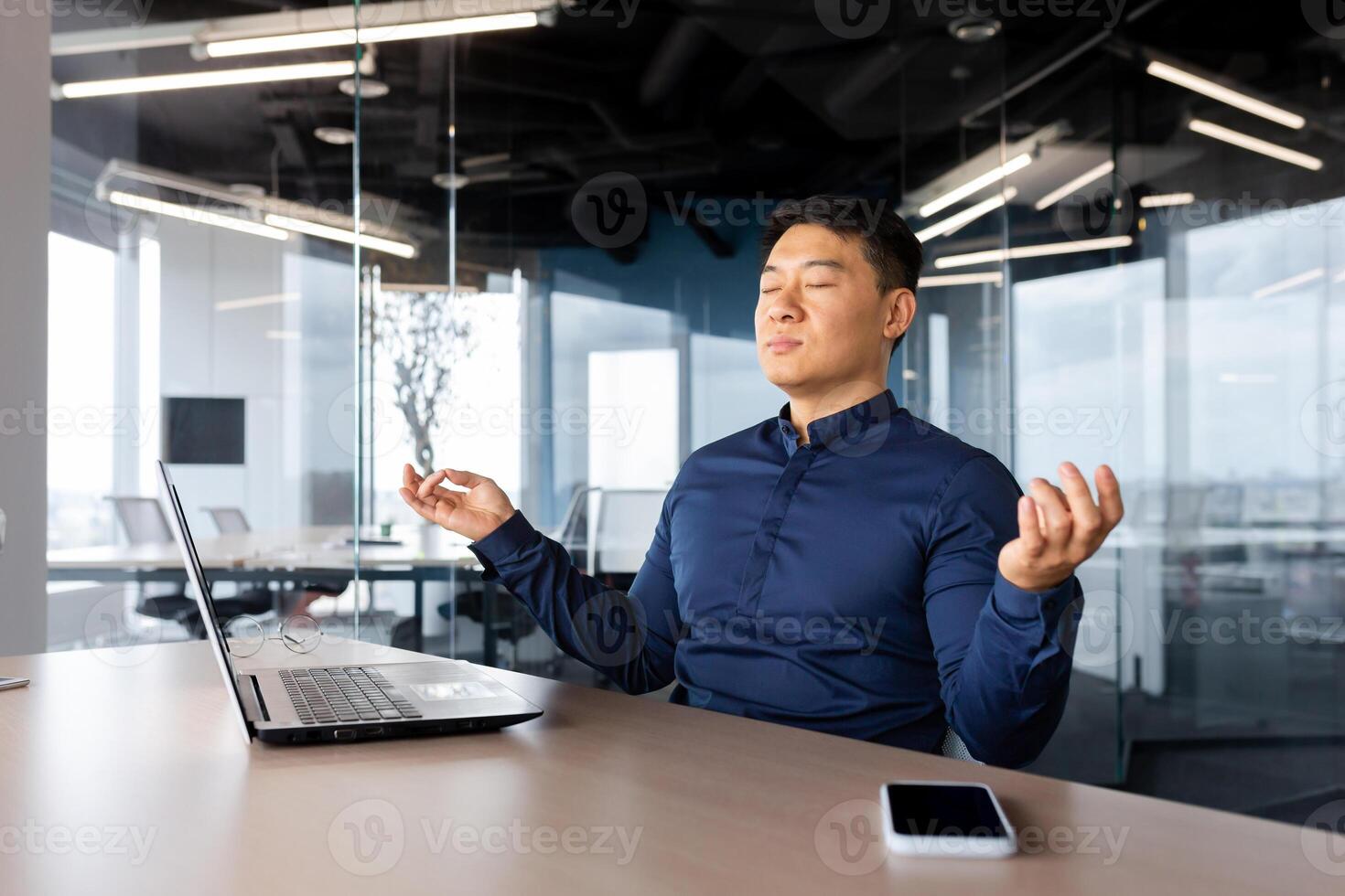 Tired young man Asian businessman meditating at work. He sits at the table in the office, took a break, closed his eyes, hands in the lotus position, relaxes. photo