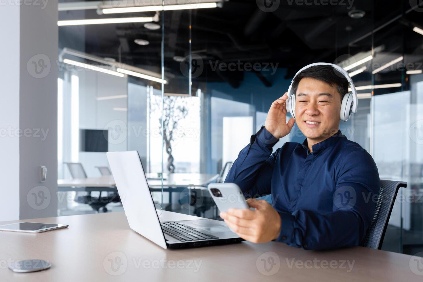 Businessman listening to music and podcast at workplace, asian man smiling and happy using laptop at work, man with headphones and phone listening to online radio inside office. photo