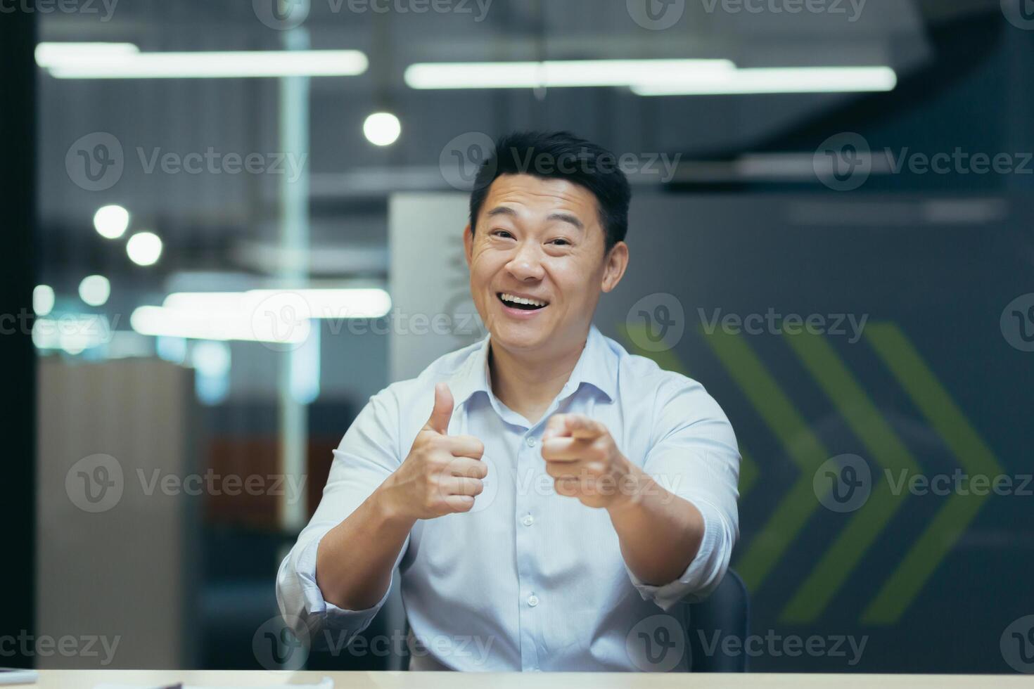 Portrait of a successful and happy Asian male businessman. He sits in the office at the work table, shows with his hands to the camera winnings, good luck, motivates, says you can do it. photo