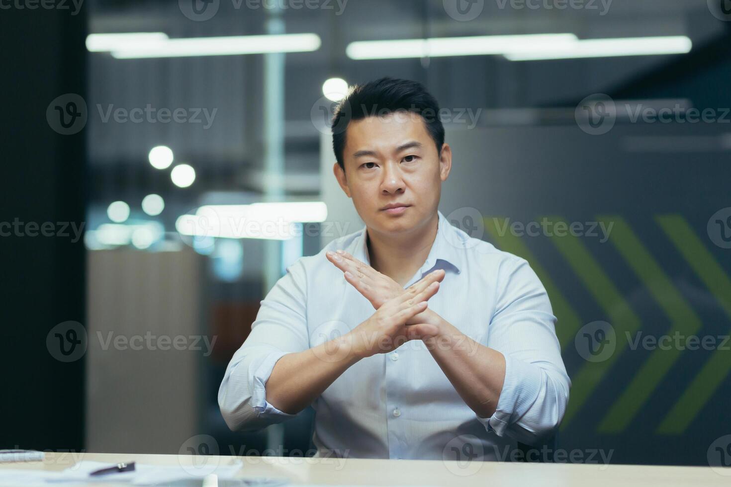 A serious young man Asian office worker sits at the desk in the office, crossed his arms in front of him, shows prostration, objection to the camera. photo