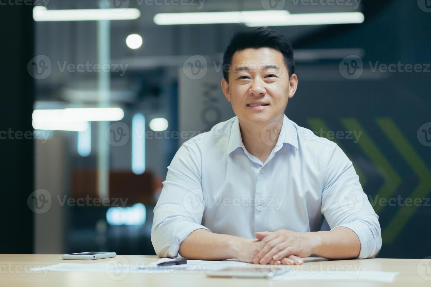 Portrait of a young Asian freelancer, designer, architect. He sits at the desk in the office, holds the phone, works with documents, looks into the camera, smilling. photo