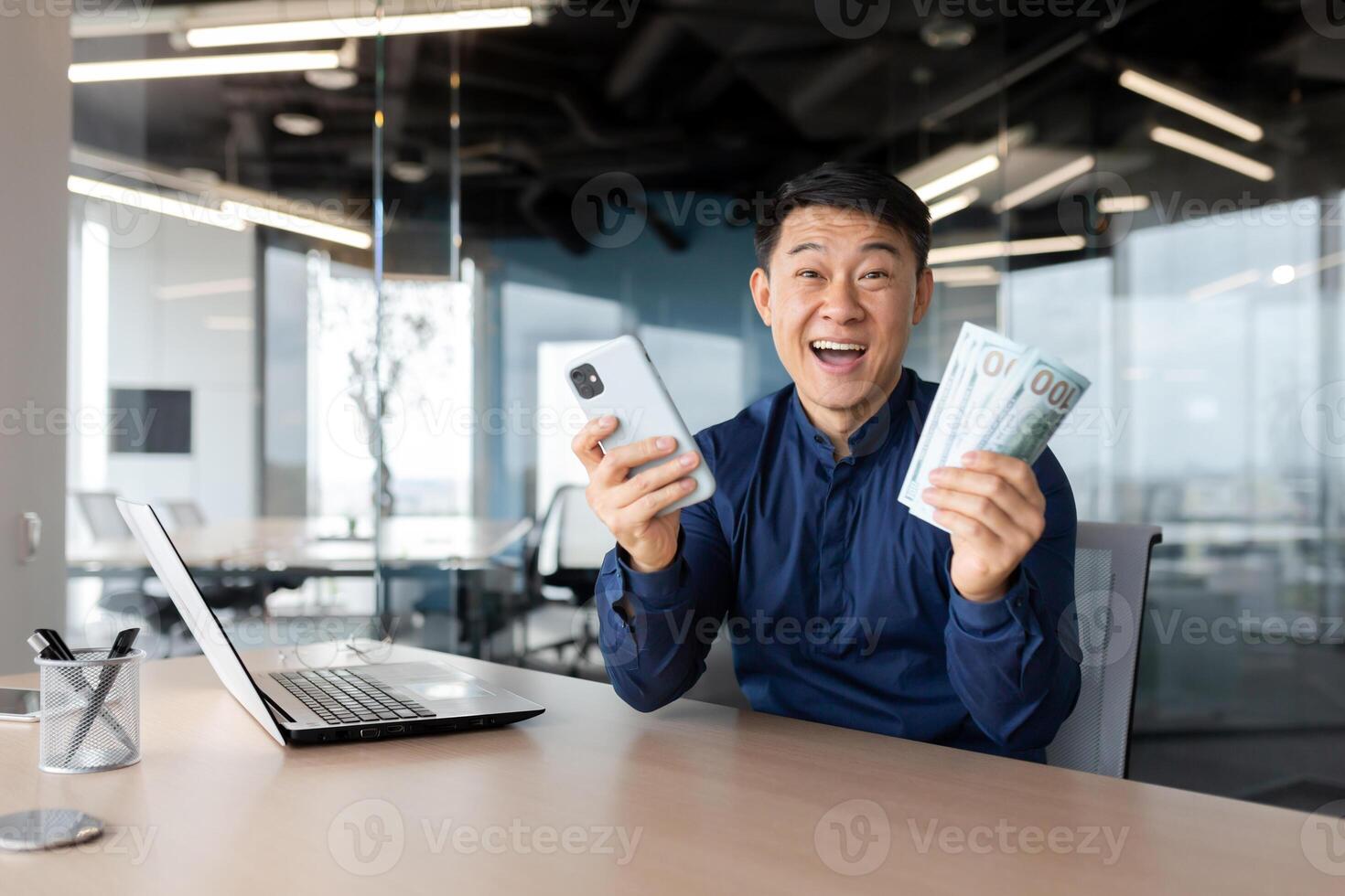 Portrait successful businessman asian smiling contentedly and looking at camera holding phone and money cash dollars in hands, celebrating victory, man in shirt working inside office using laptop. photo