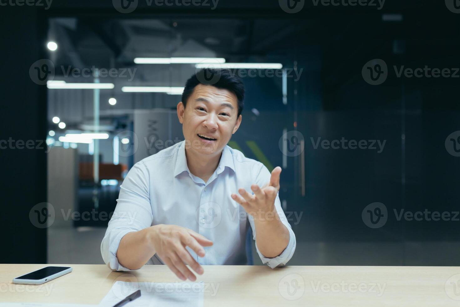 Successful and happy Asian businessman looking at camera, talking on call, cheerfully gesticulating to interlocutor, online meeting, man working in office at work photo