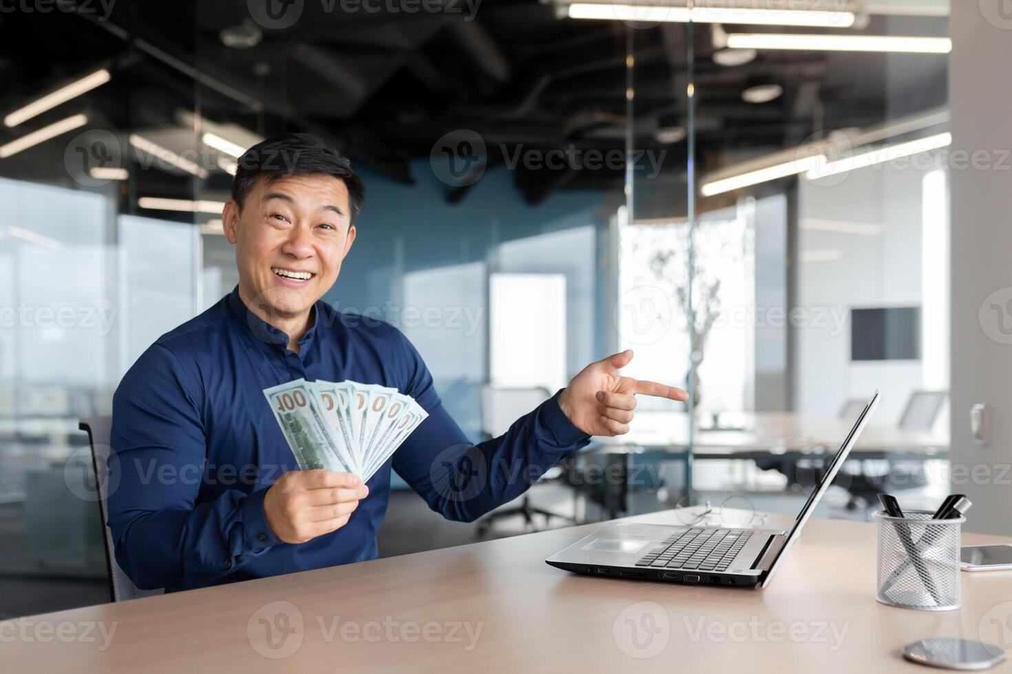 Happy young man Asian businessman sitting in the office holding cash money in his hands. He works at a laptop, points his finger at the monitor, has online meeting with partners, investors, smiles. photo