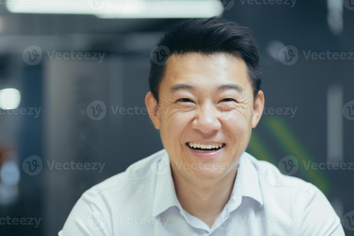 Close-up photo. Portrait of a handsome young Asian businessman. He is sitting in the office in a white shirt, looking at the camera, smiling. photo