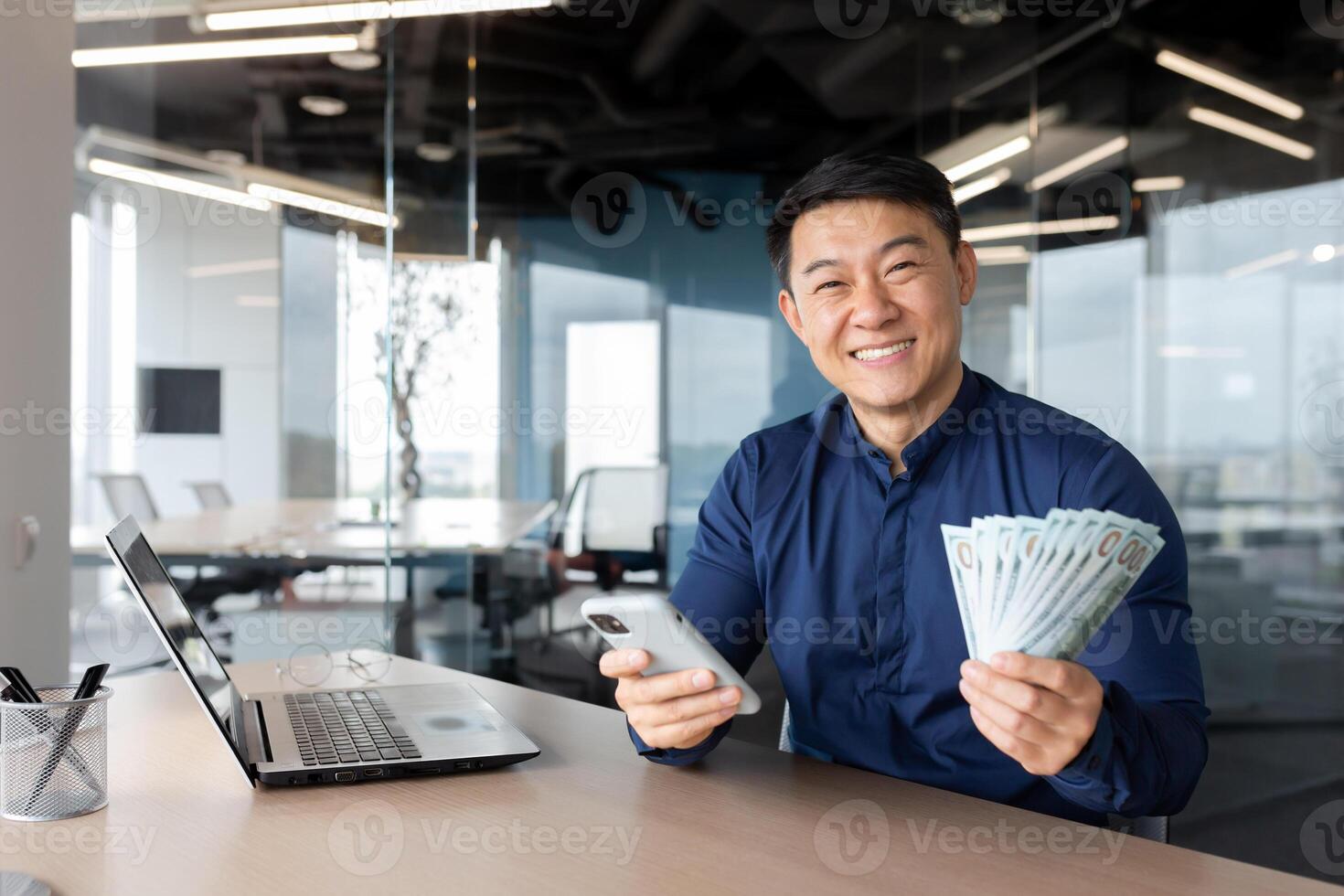 A young Asian man is sitting in the office. He is holding a phone and cash money. He looks at the camera, smiles. photo