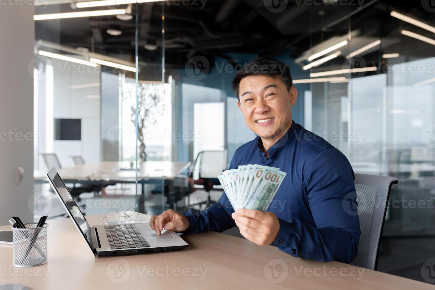 A young Asian man sits in the office at a table with a laptop. He holds a phone and cash in his hands, points to the camera, smiles. photo