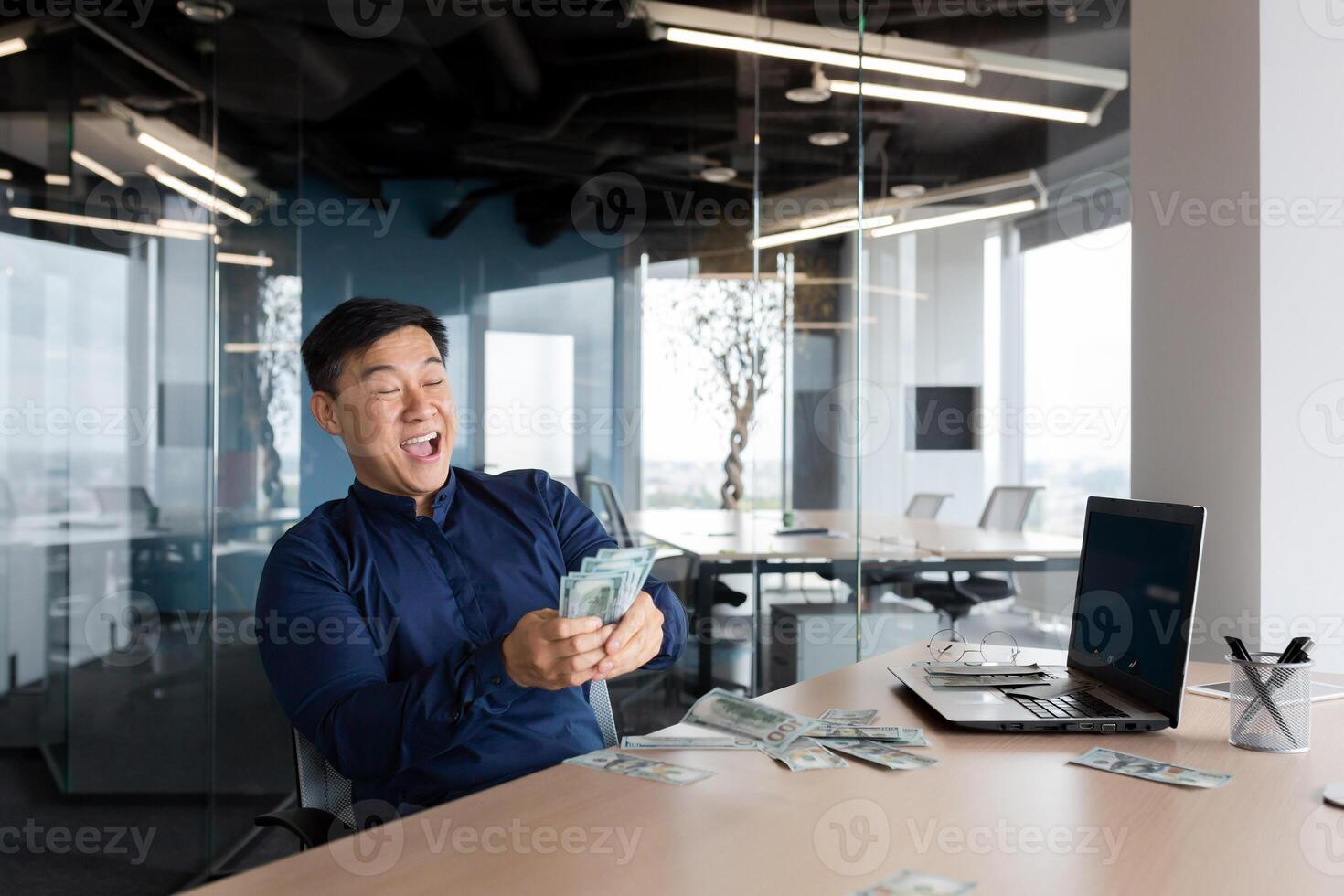 A happy young Asian man, an office worker, an entrepreneur holds cash money in his hands, received a salary, a bonus. He is sitting at a table in the office, rejoices, shouts with joy. photo
