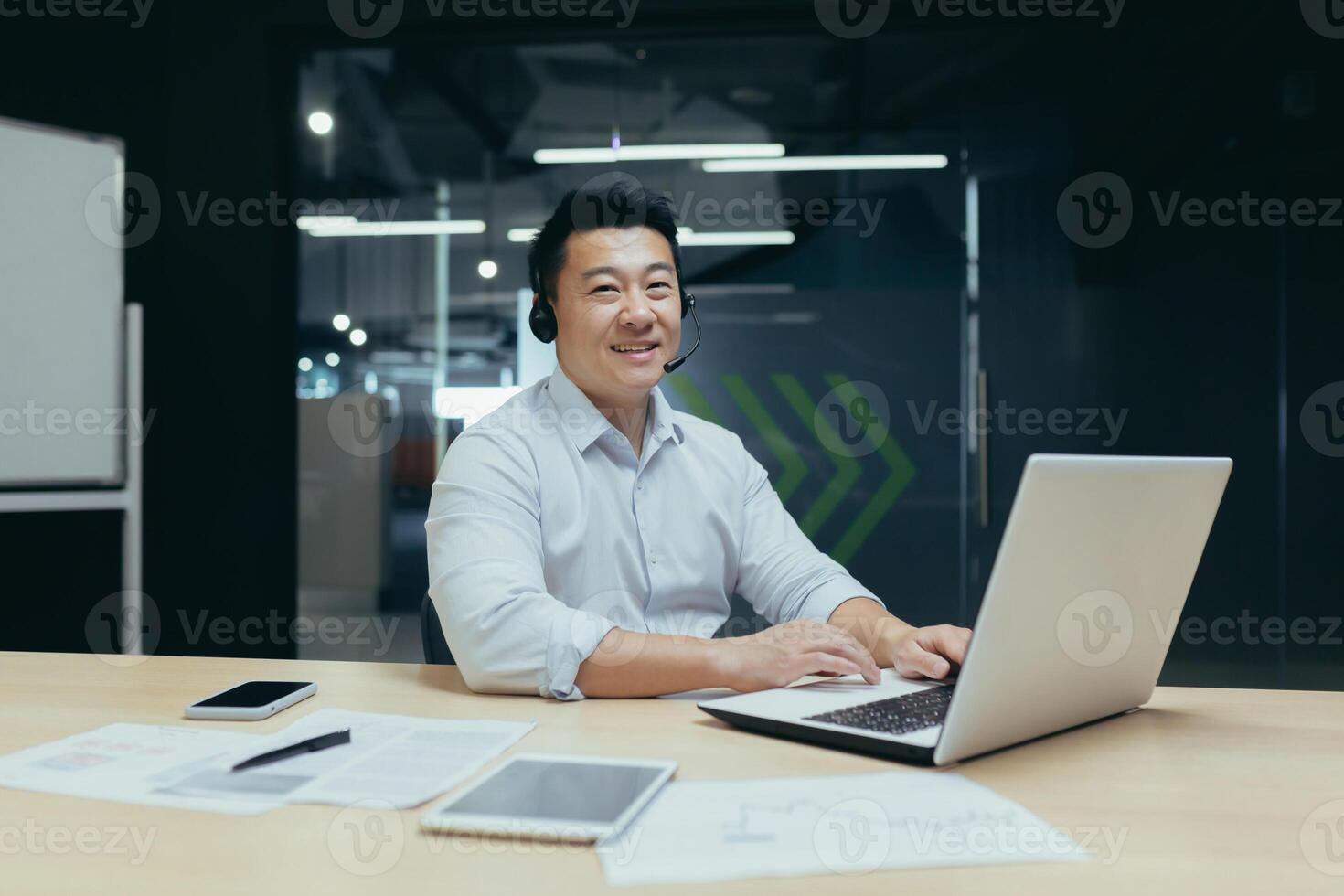 Portrait of a young Asian man sitting in the office at a desk in a headset, working on a laptop. He looks at the camera, smiles, points with his finger super. photo