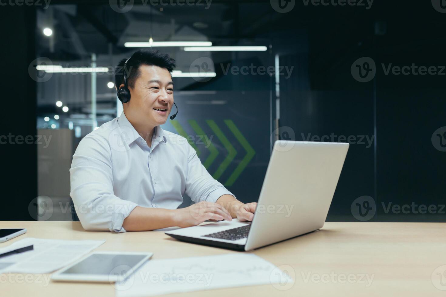 A young handsome Asian businessman in headphones with a microphone sits at a laptop in the office, conducts an online conference, a remote meeting with clients, partners. Tells, explains, smiles photo
